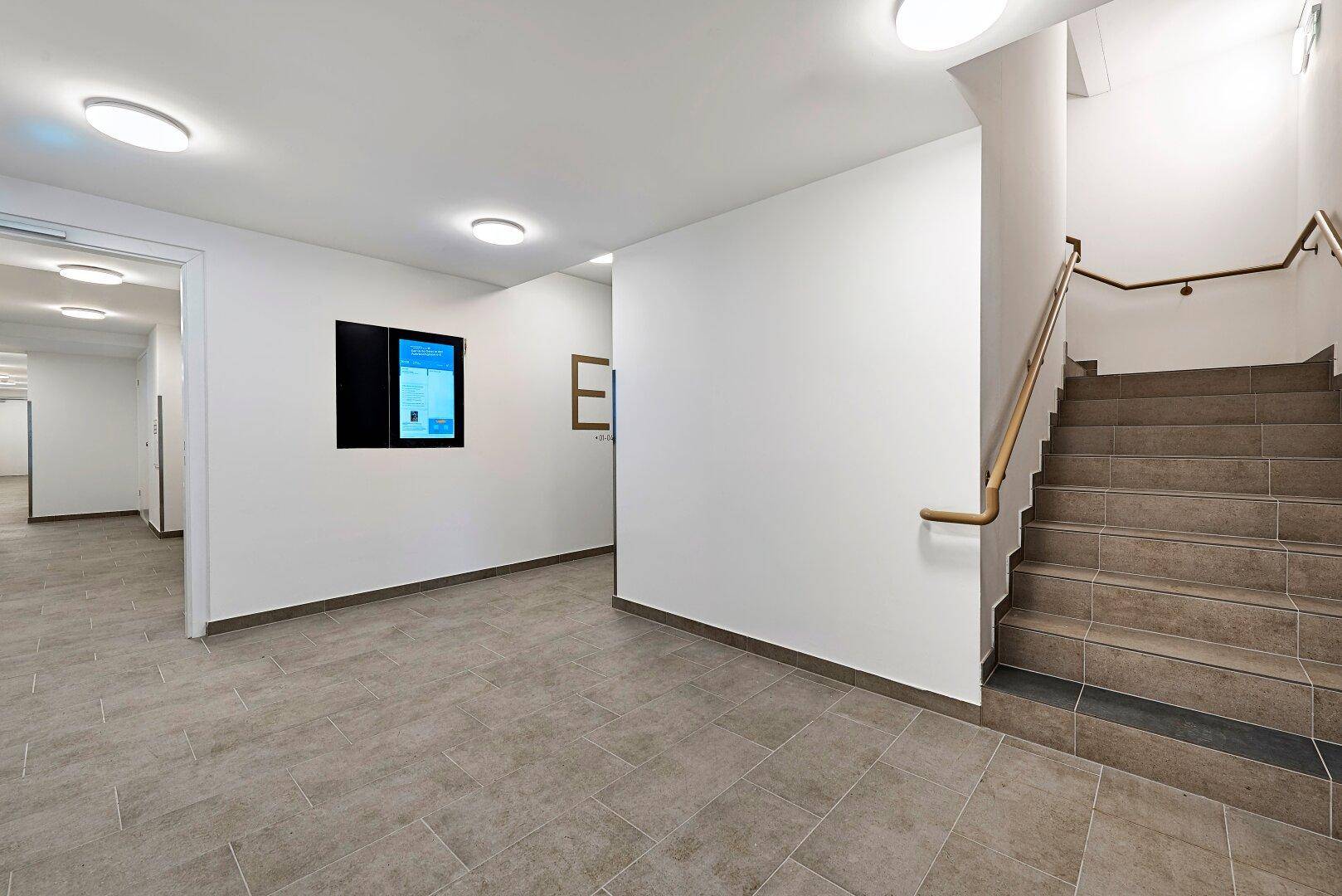 Heller Eingangsbereich mit modernem Fliesenboden, weiß gestrichenen Wänden und einer geräumigen Treppe mit Holzgeländer.