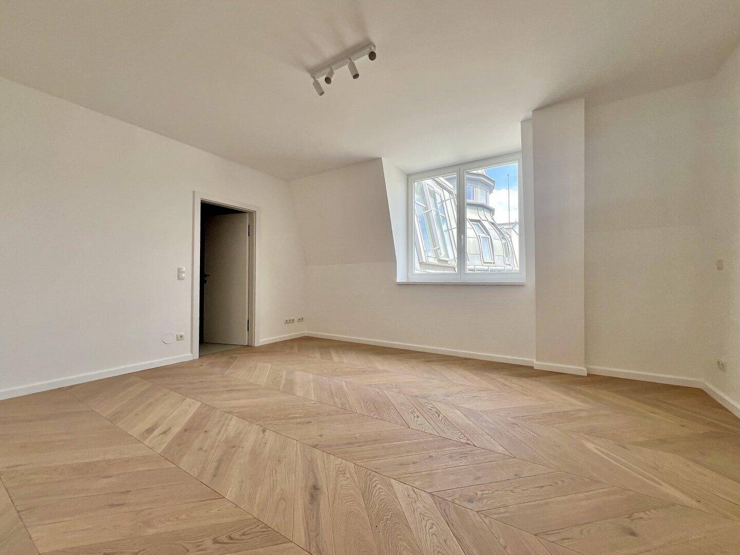 Helles Zimmer mit Fischgrätparkett und einem Fenster mit Blick auf die Dächer der Stadt.