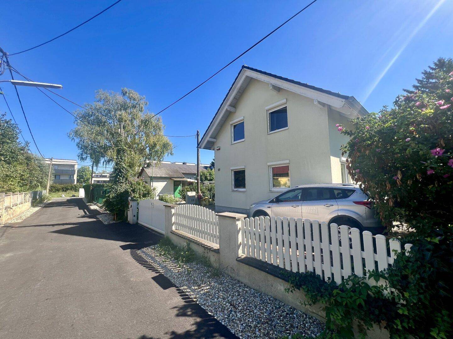 Einfamilienhaus mit heller Fassade, weißem Zaun und einem geparkten Auto vor dem Eingang.