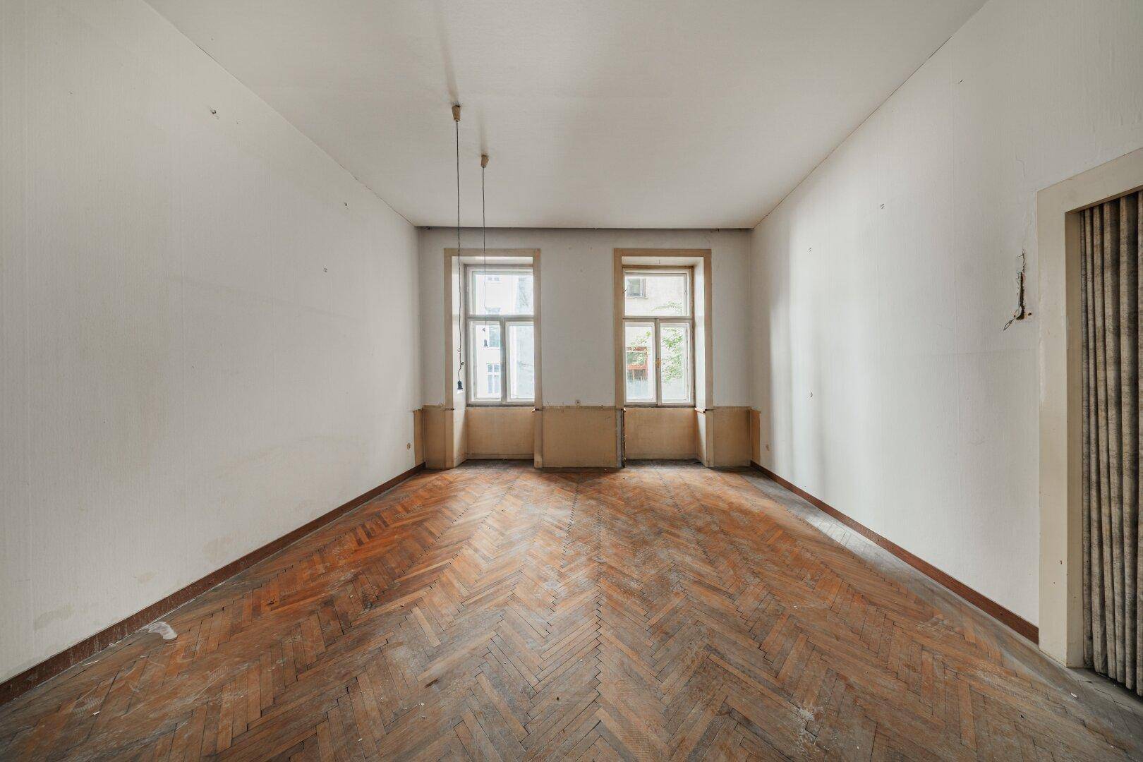 Leerer Raum mit Fischgrätparkett und zwei hohen Fenstern, bereit zur Renovierung.