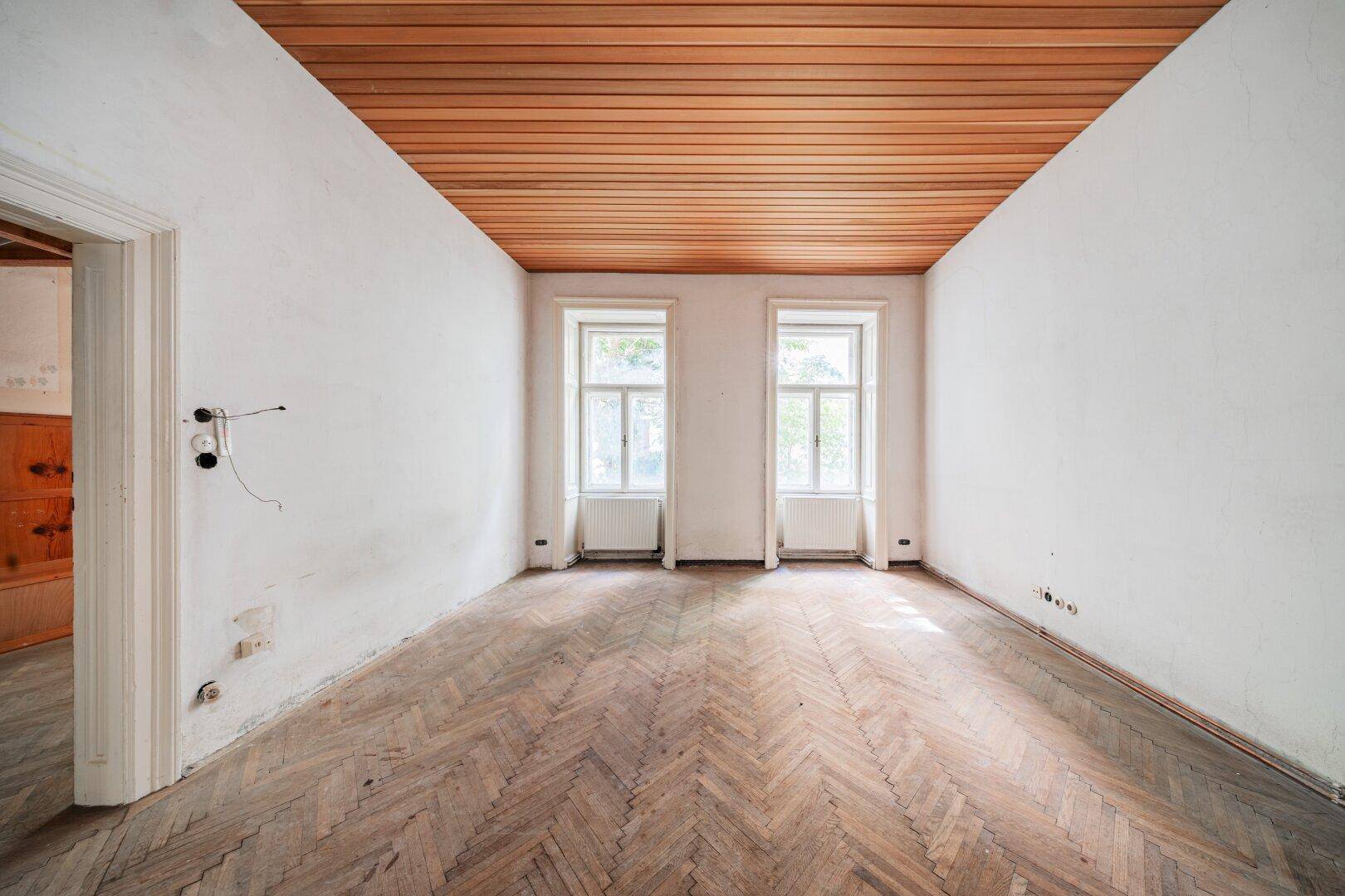 Leerer Raum mit Fischgrätparkett, hohen Decken und zwei Fenstern, bereit für die Renovierung.