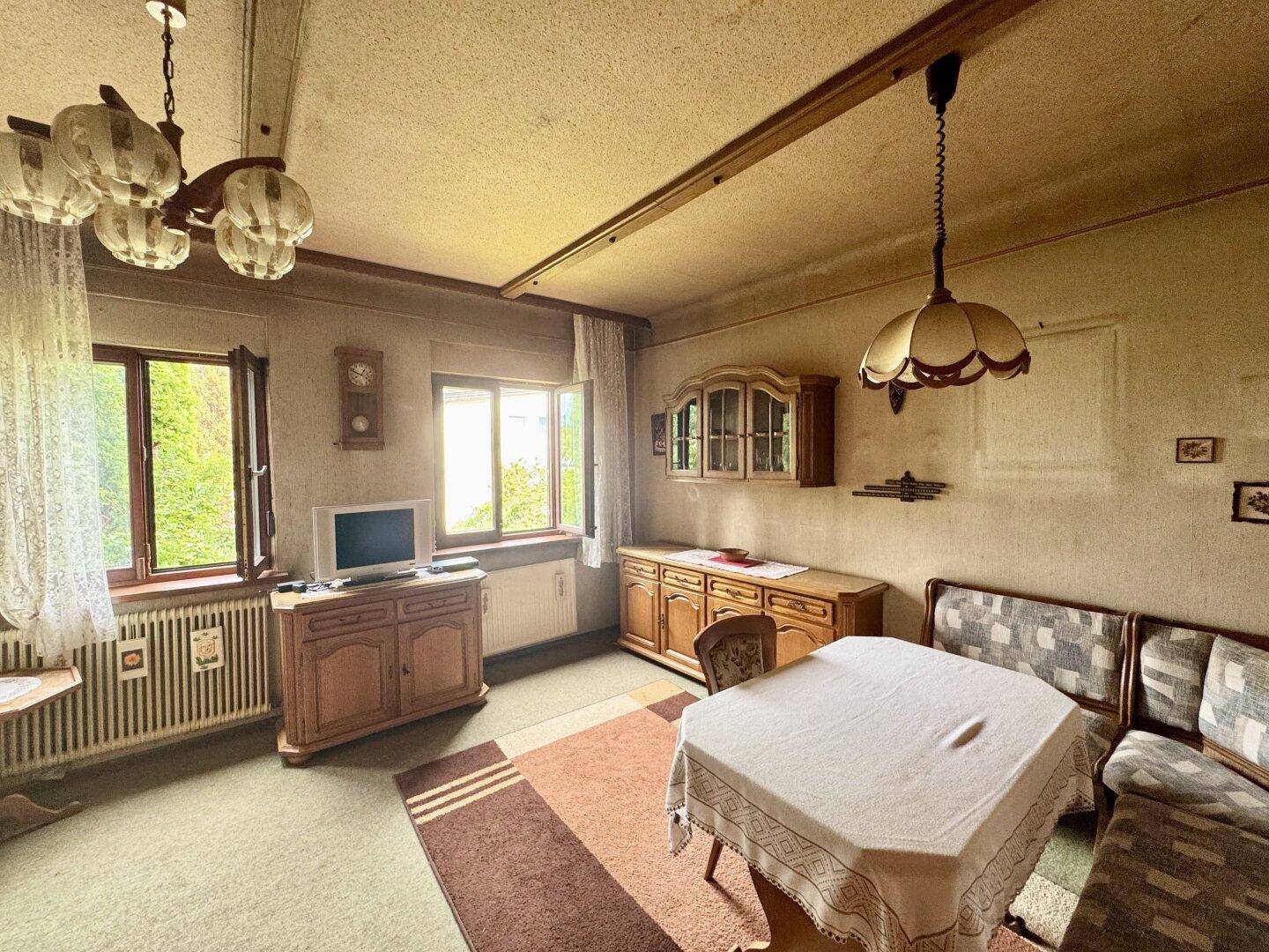 Traditioneller Wohn-Essbereich mit Teppichboden, Holzmöbeln und Fenstern mit Blick ins Grüne.