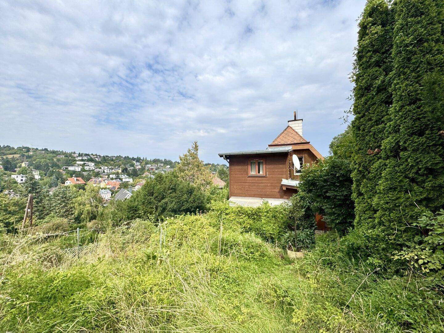 Seitenansicht eines Holzhauses mit Satteldach, eingebettet in eine grüne Landschaft mit Fernblick auf eine Siedlung.