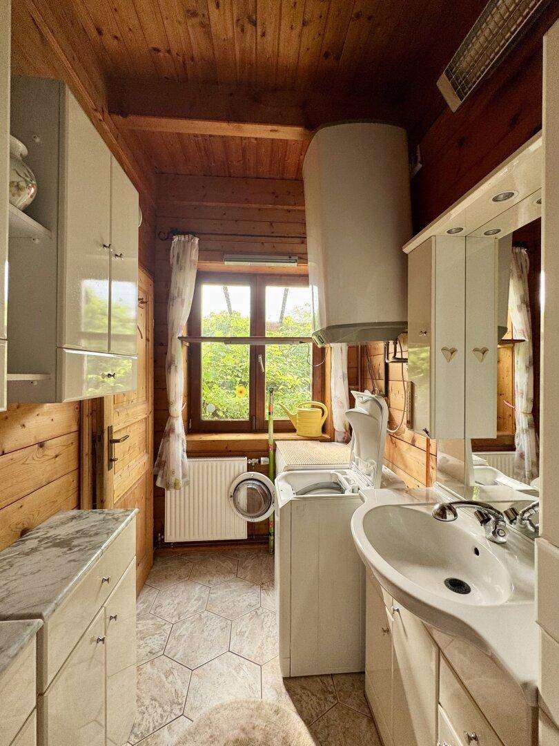 Funktionaler Waschraum mit Fliesenboden, Holzwänden, Waschmaschine und Fenster mit Blick ins Grüne.
