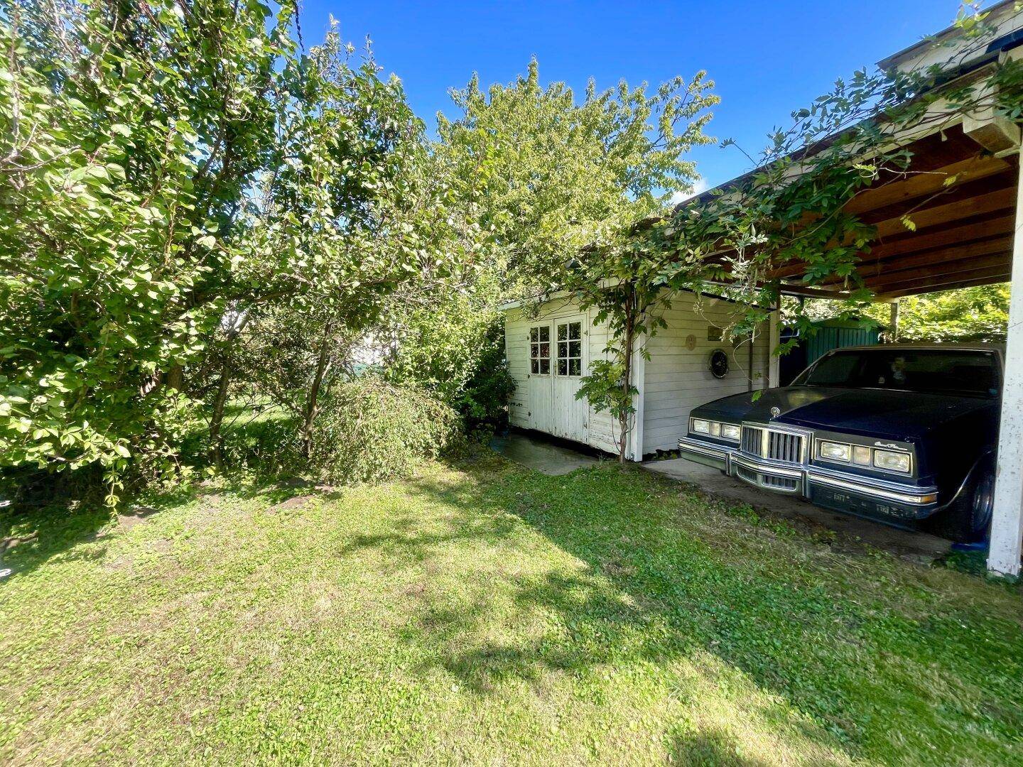 Garten mit Carport und einem kleinen weißen Gartenhaus, umgeben von Bäumen.