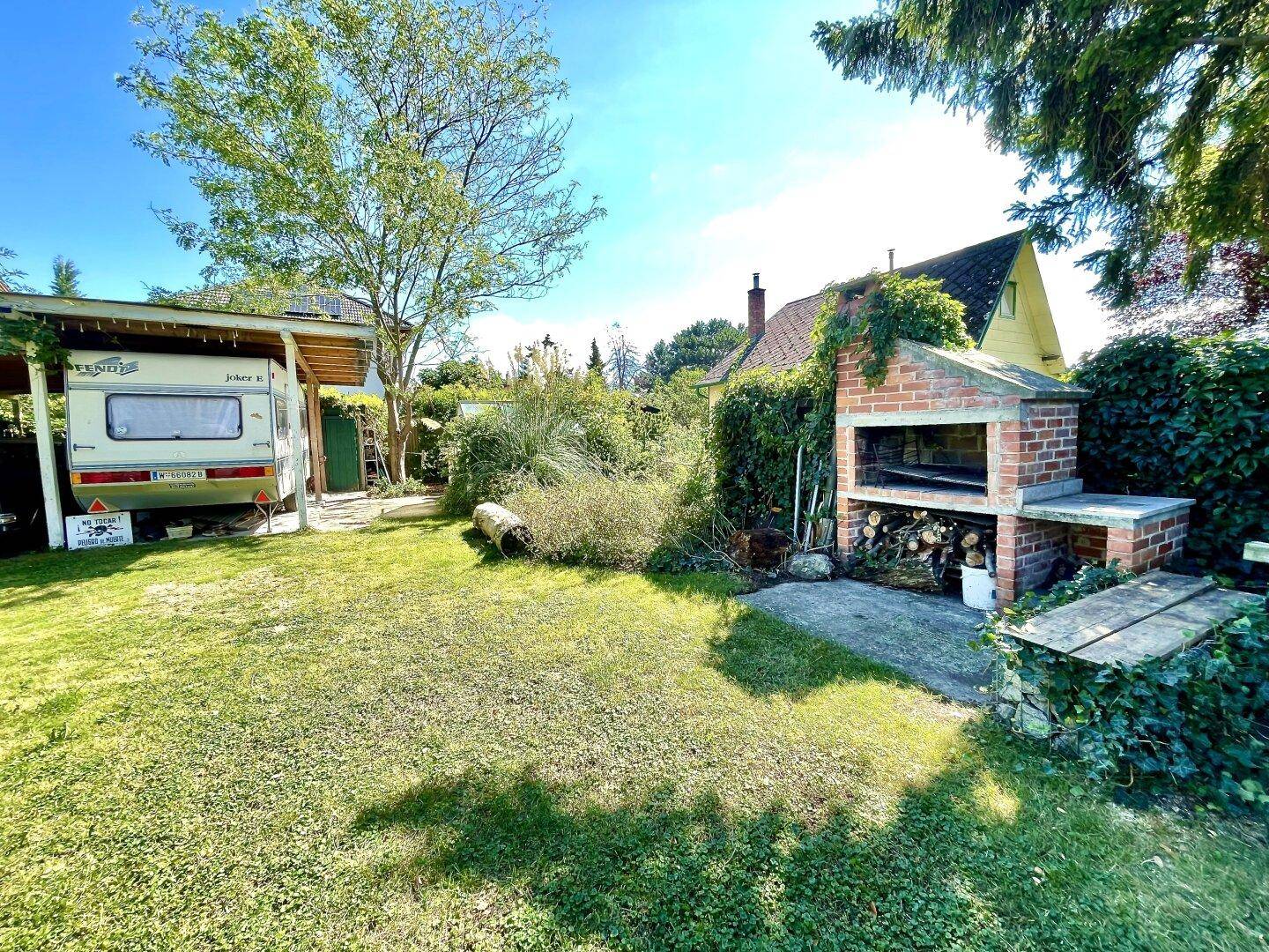 Gartenbereich mit gemauertem Grillplatz und Holzschuppen, umgeben von üppigem Grün.
