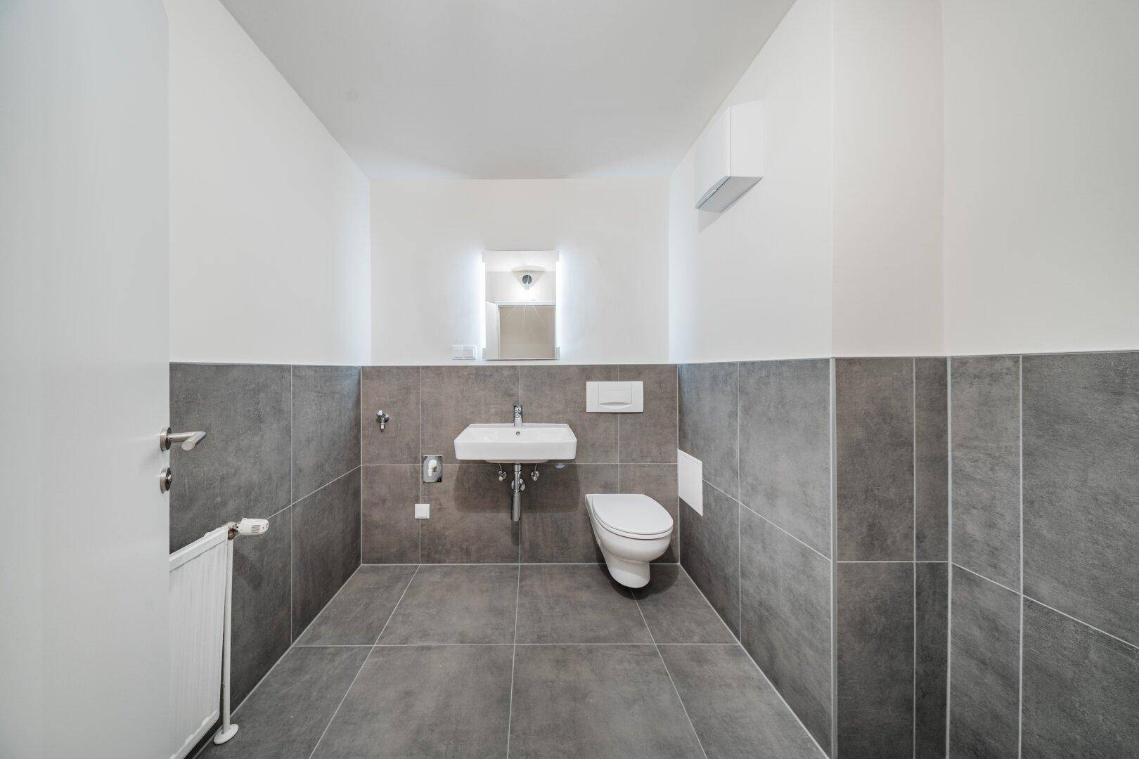 Modernes Gäste-WC mit grauen Fliesen, wandhängendem WC und kleinem Waschbecken.