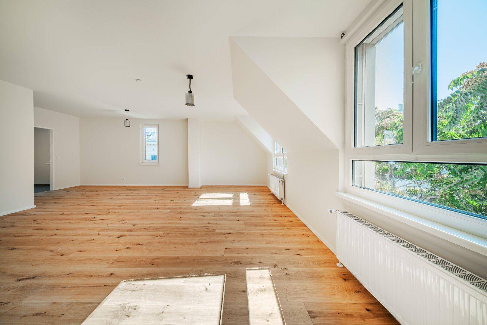 Großes Dachgeschosszimmer mit Holzboden, Dachschrägen und hellen Fenstern mit Blick ins Grüne.