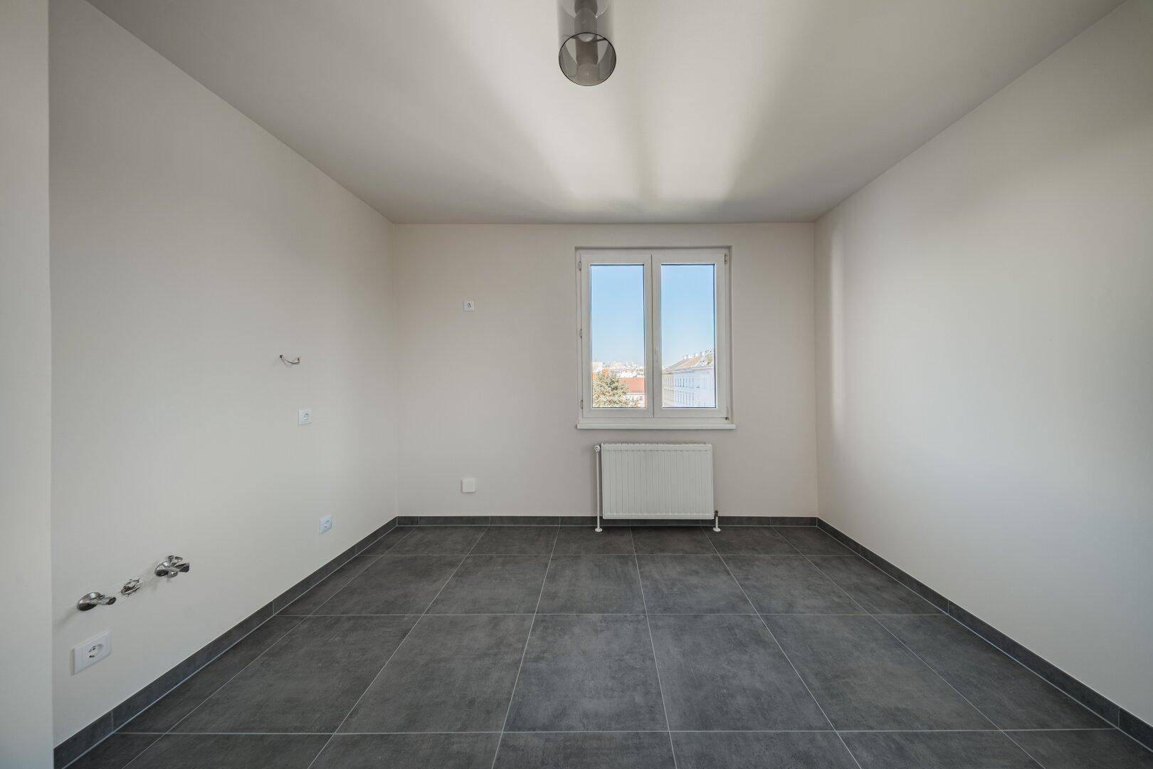 Kleiner Raum mit grauen Fliesen, Fenster und Anschlüssen, ideal als Abstell- oder Hauswirtschaftsraum.