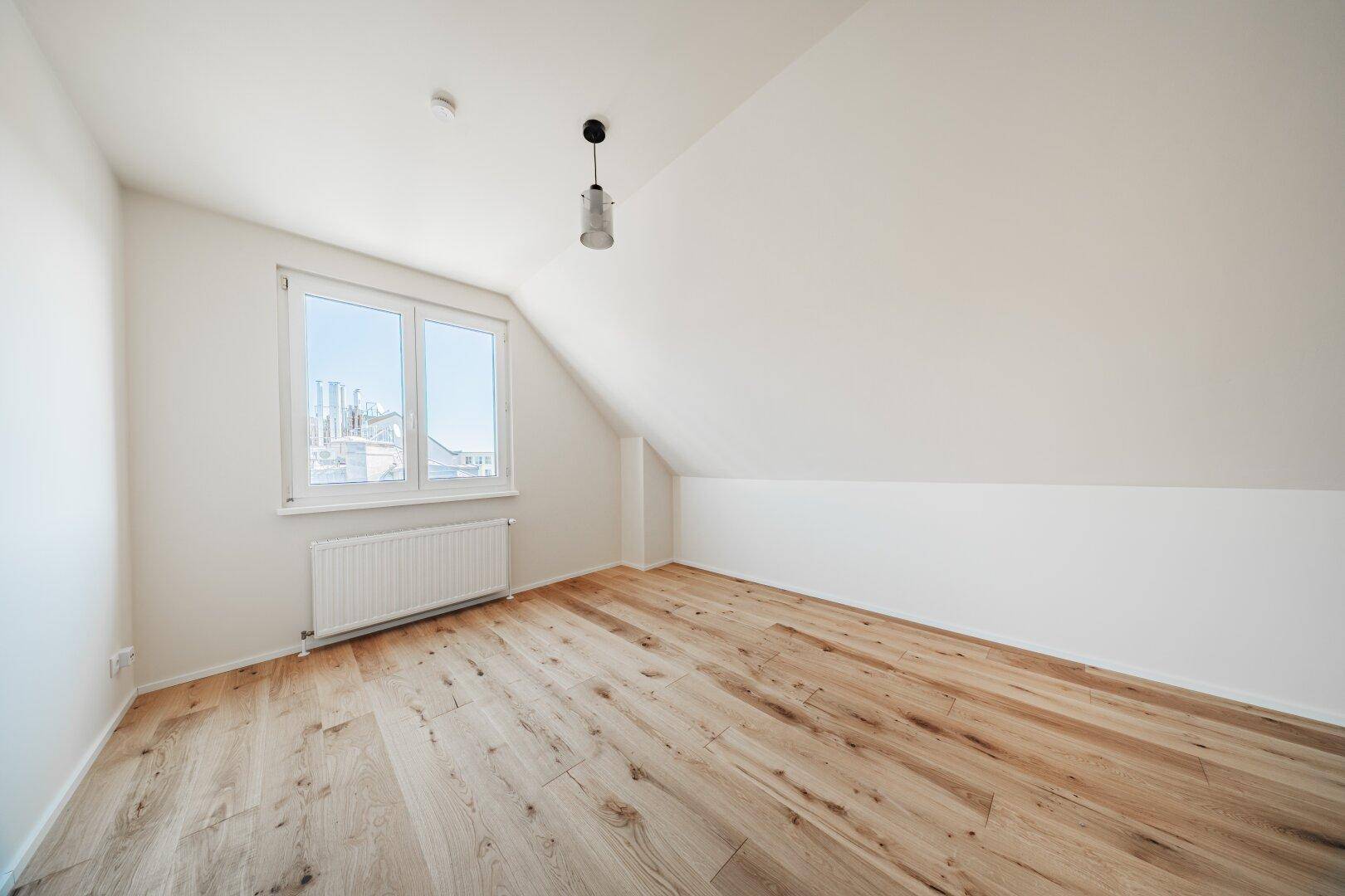 Helles Dachgeschosszimmer mit Holzboden, Dachschräge und Fenster mit Stadtblick.