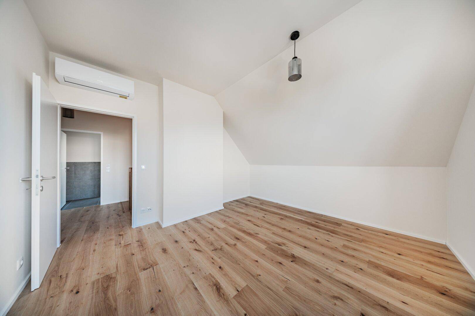Dachgeschosszimmer mit Holzboden, Dachschräge und Klimaanlage, mit Zugang zu einem weiteren Raum.