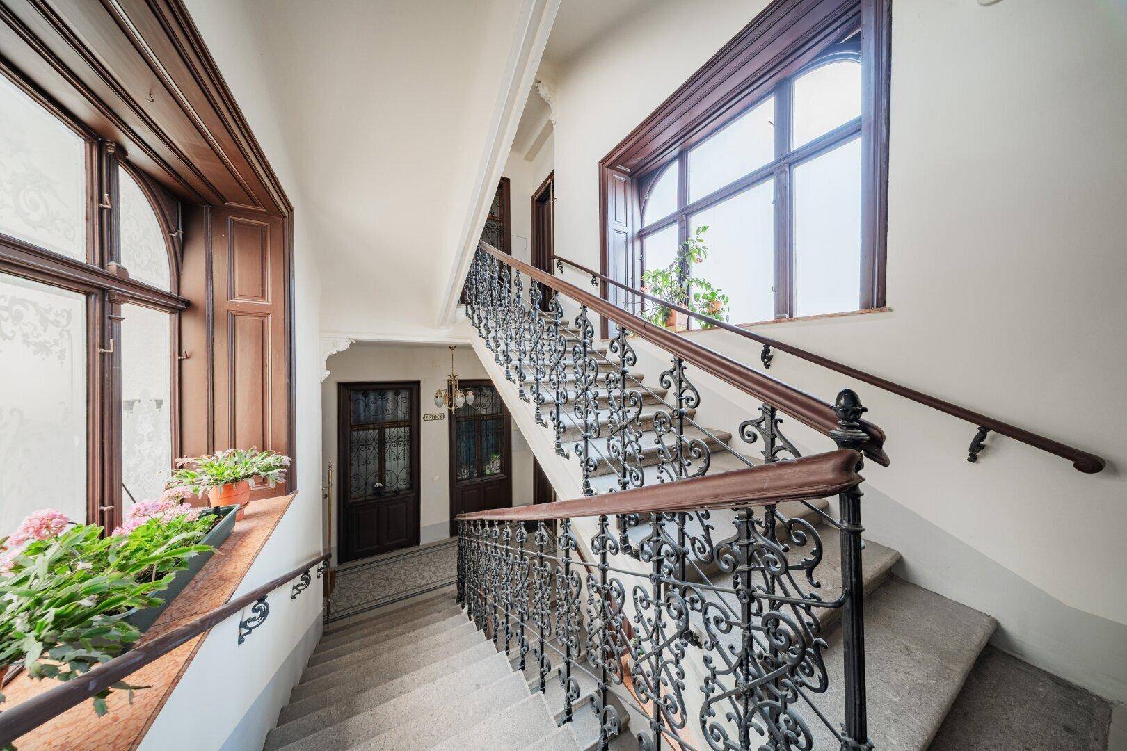 Elegantes Treppenhaus in einem Altbau mit schmiedeeisernem Geländer, großen Fenstern und Pflanzen.
