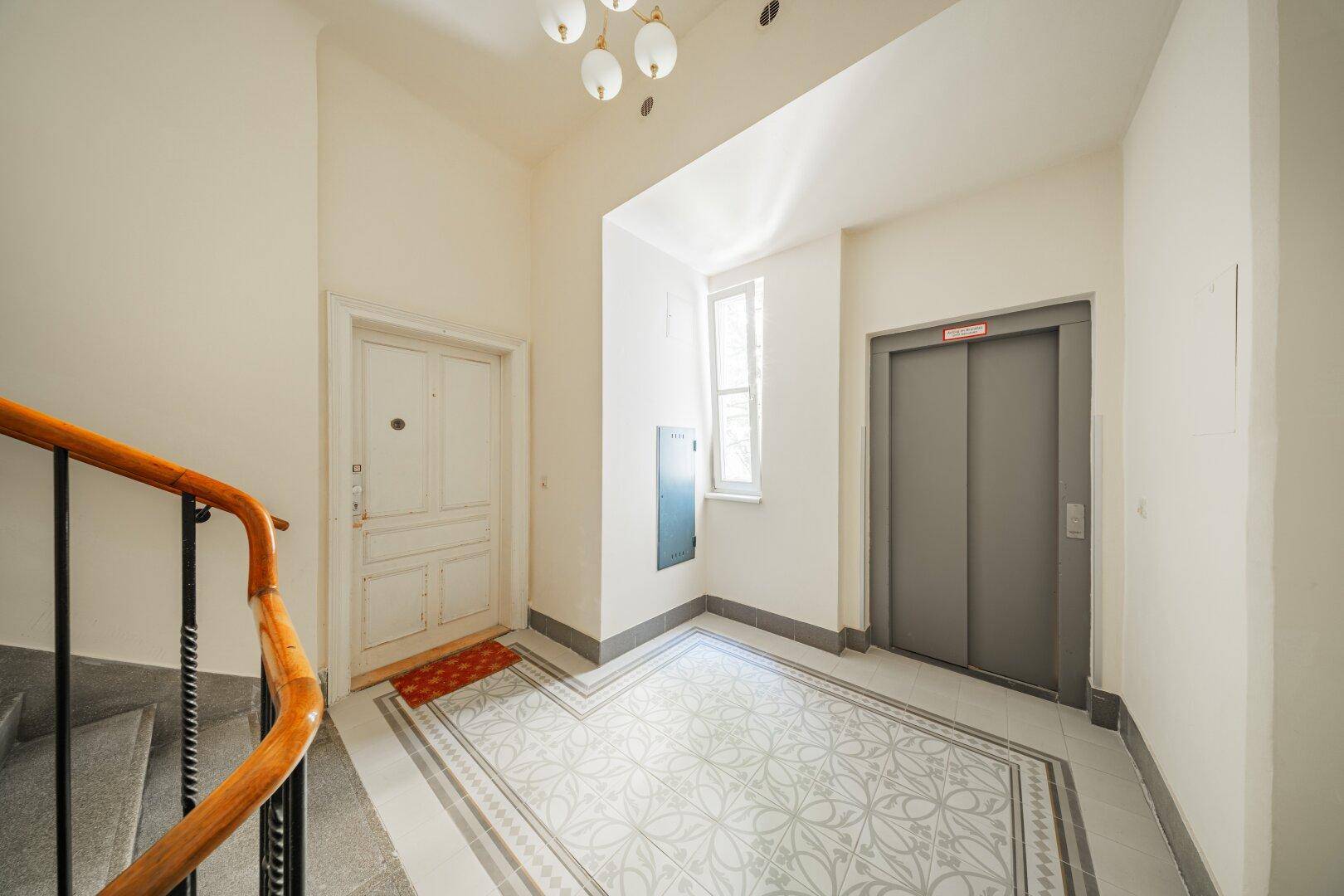 Heller Eingangsbereich mit Aufzug, Treppenhaus und dekorativen Bodenfliesen in einem Altbau.