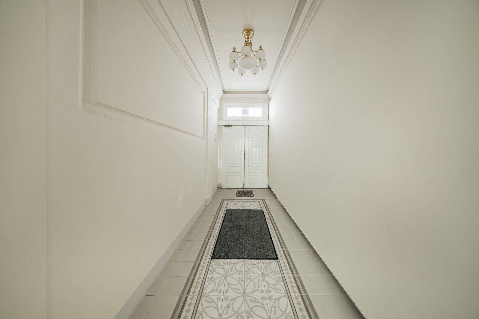 Schmaler Flur mit hohen Decken, klassischer Beleuchtung und gemustertem Fliesenboden in einem Altbau.