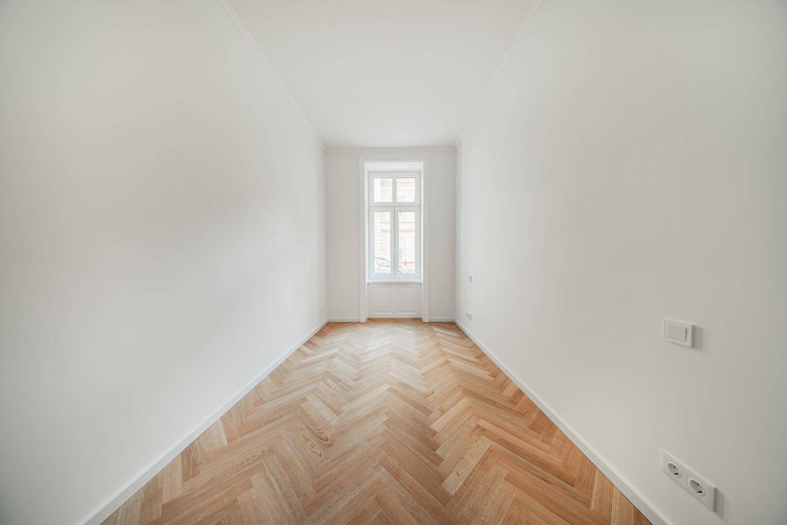 Helles, leeres Zimmer mit Fischgrätparkett und einem großen Fenster, ideal als Schlafzimmer.