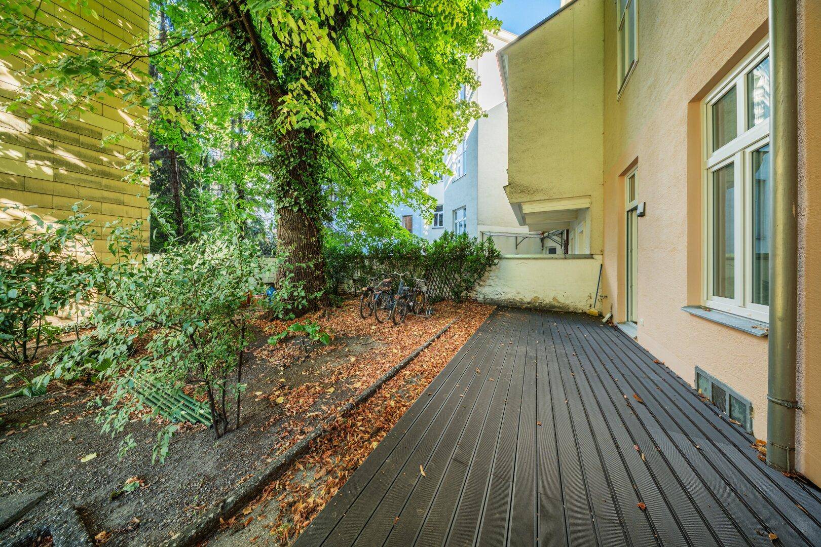 Dunkle Holzterrasse mit altem Baum und Fahrradstellplatz im grünen Außenbereich.