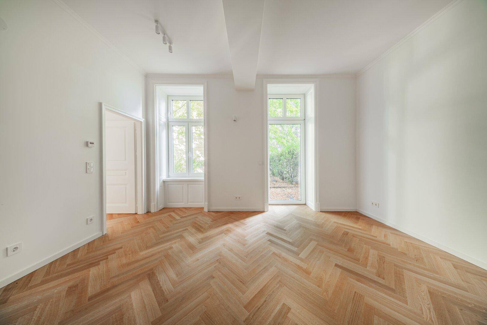 Geräumiges Zimmer mit elegantem Fischgrätparkett und zwei großen Fenstern zum Garten.