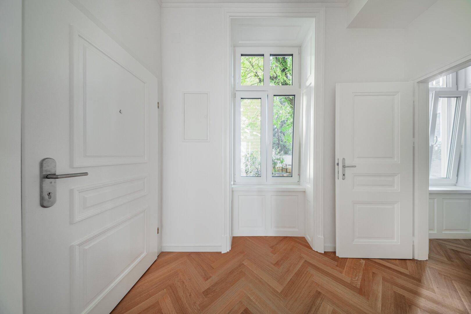 Heller Flur mit Fischgrätparkett, weißen Türen und einem hohen Fenster mit Blick ins Grüne.