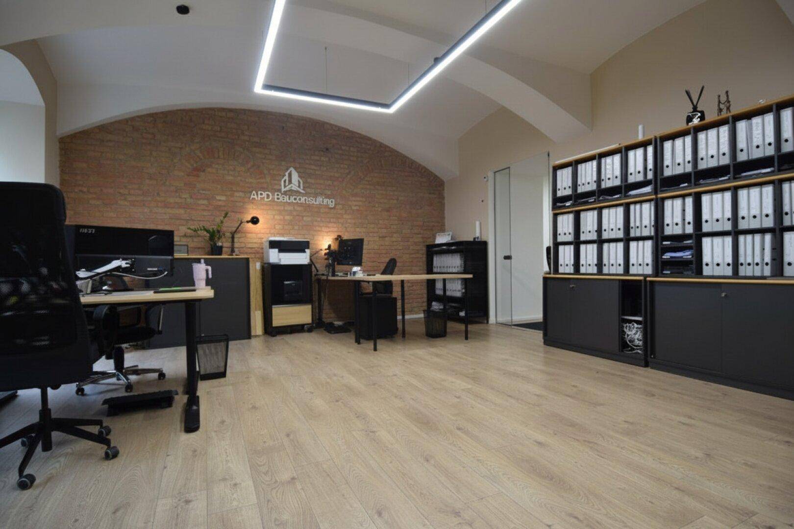 Modernes Büro mit freigelegter Ziegelwand, Holzboden und effizienter Deckenbeleuchtung.