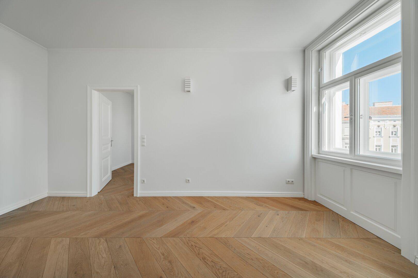 Heller Raum mit klassischem Fischgrätparkett und großen Fenstern, die viel Tageslicht hereinlassen.