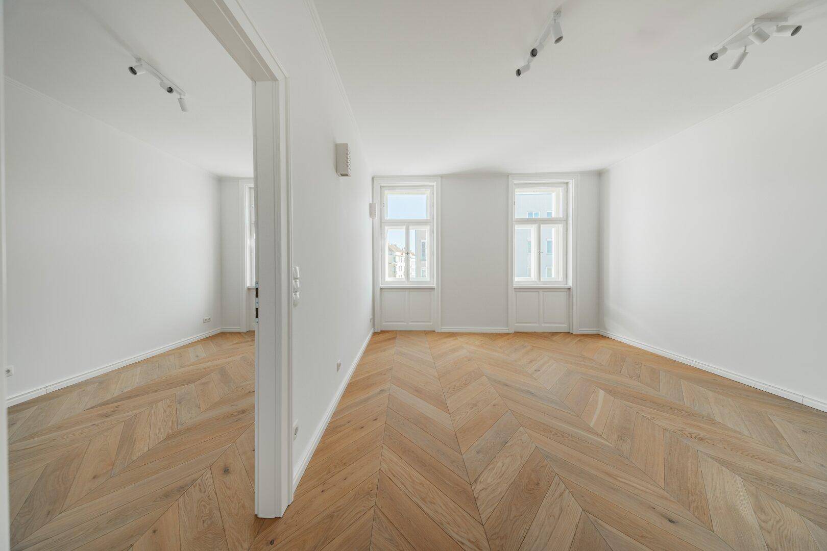 Geräumiges, lichtdurchflutetes Zimmer mit elegantem Fischgrätparkett und hohen Decken.