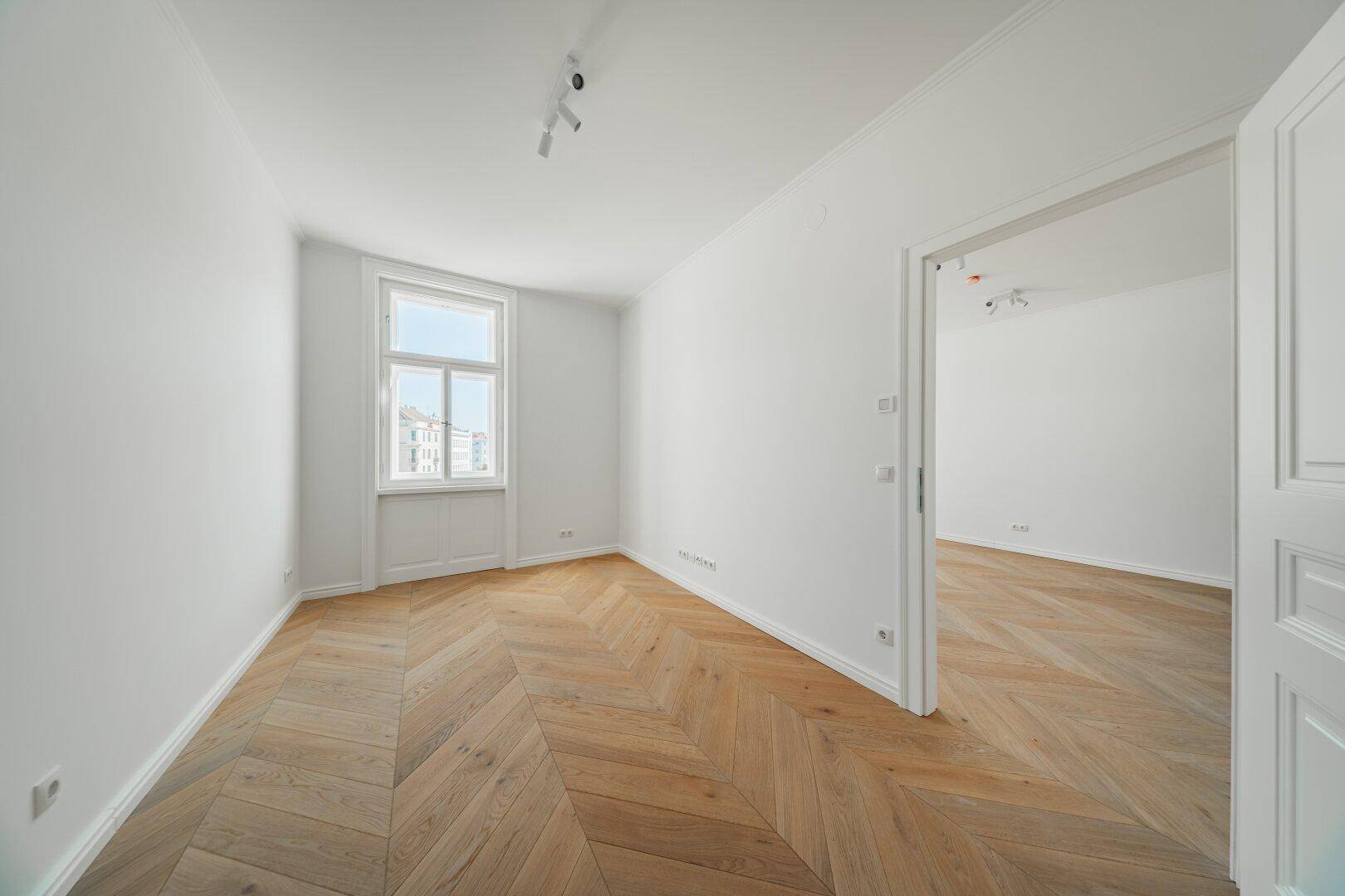 Heller Wohnraum mit Fischgrätparkett und Zugang zu einem weiteren Zimmer, ideal für flexible Nutzung.
