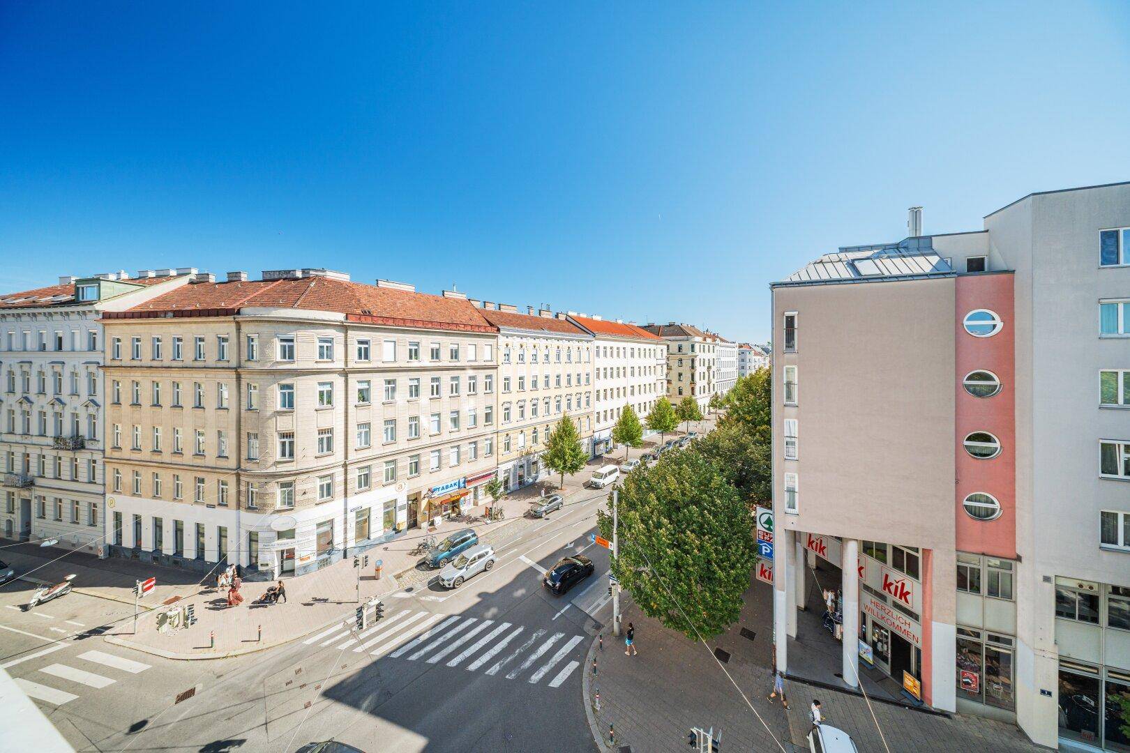 Weitläufige Stadtansicht mit belebter Straße, historischen Gebäuden und Geschäften unter blauem Himmel.