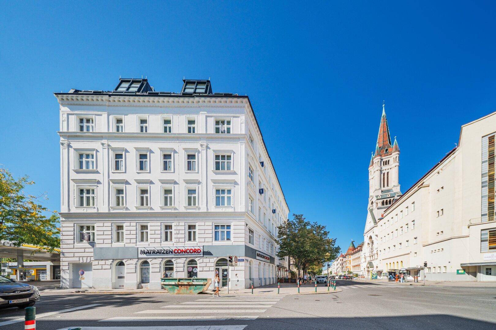 Historisches Eckgebäude mit weißer Fassade und einem Kirchturm im Hintergrund unter blauem Himmel.