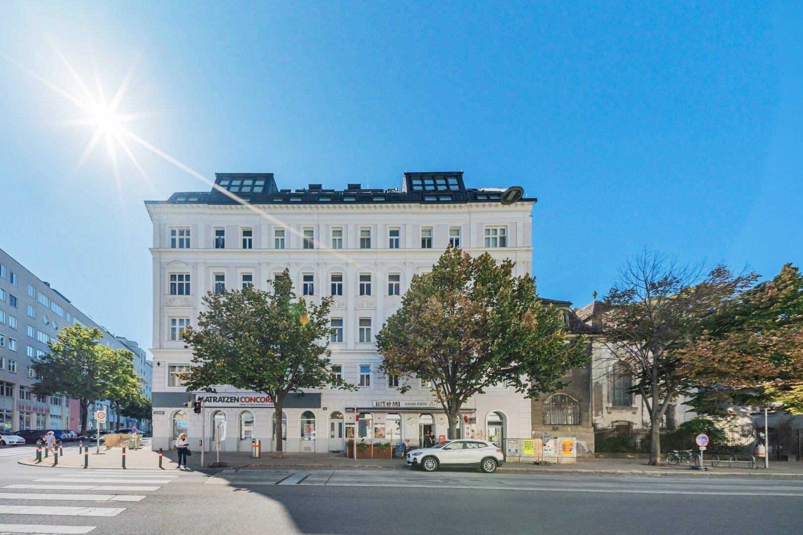 Sonnenbeschienenes Eckgebäude mit weißer Fassade und Geschäften im Erdgeschoss an einem klaren Tag.