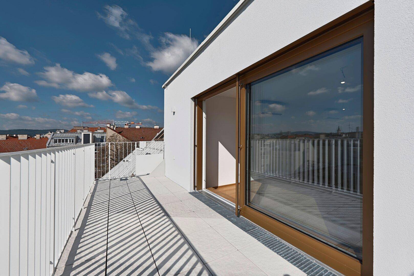 Langer, sonniger Balkon mit weißem Geländer und Blick über die Stadt.