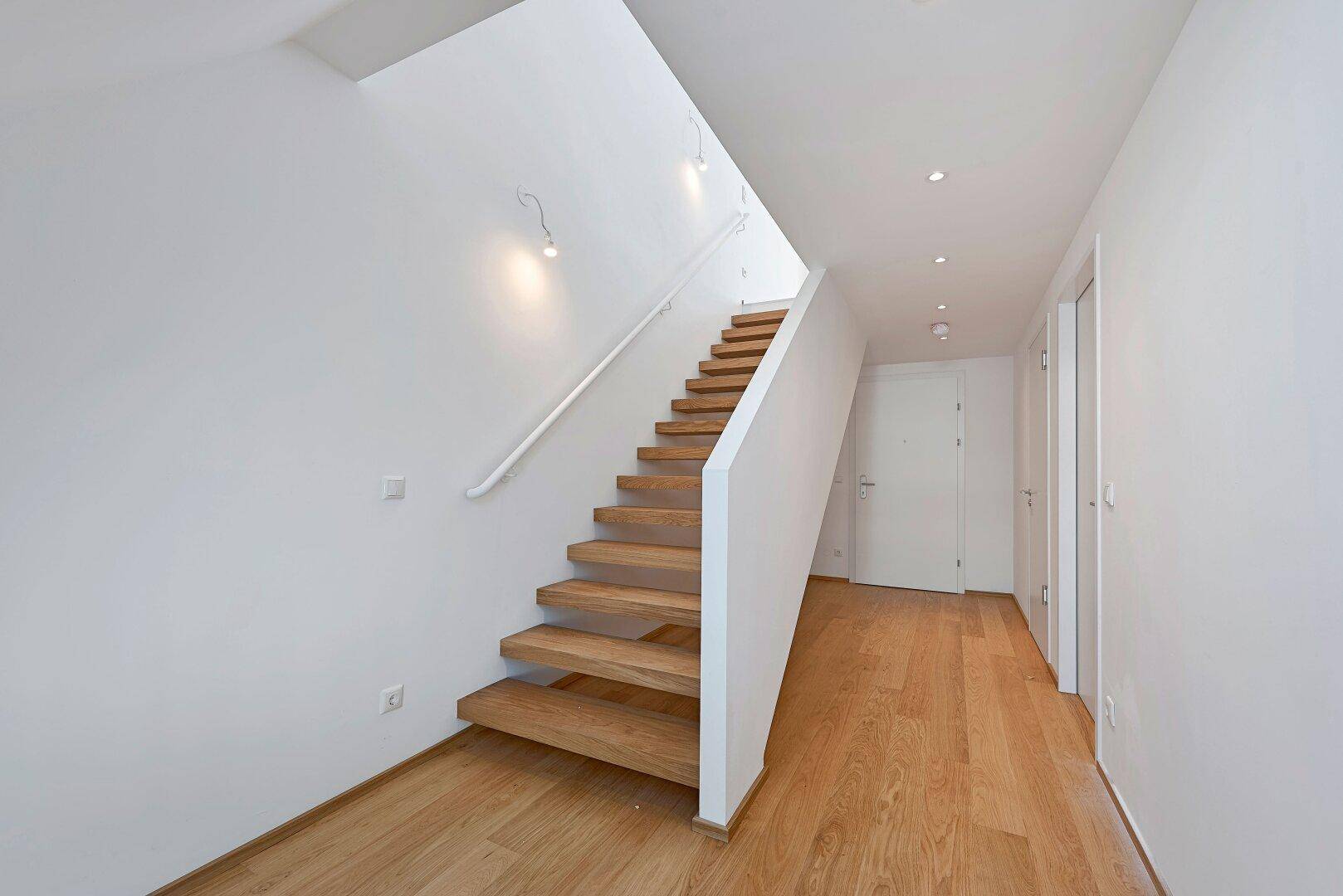 Heller Flur mit moderner Holztreppe, weißem Geländer und integrierter Deckenbeleuchtung.