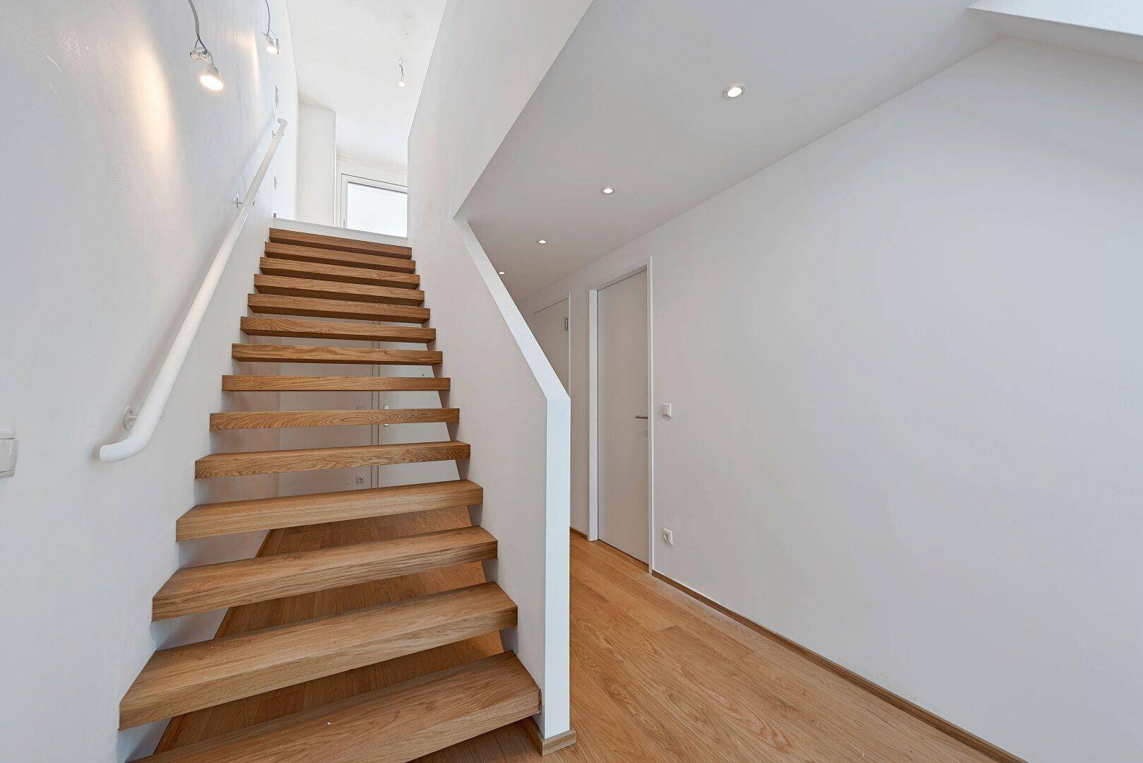 Blick auf eine moderne Holztreppe mit weißem Geländer und minimalistischer Beleuchtung im Flurbereich.