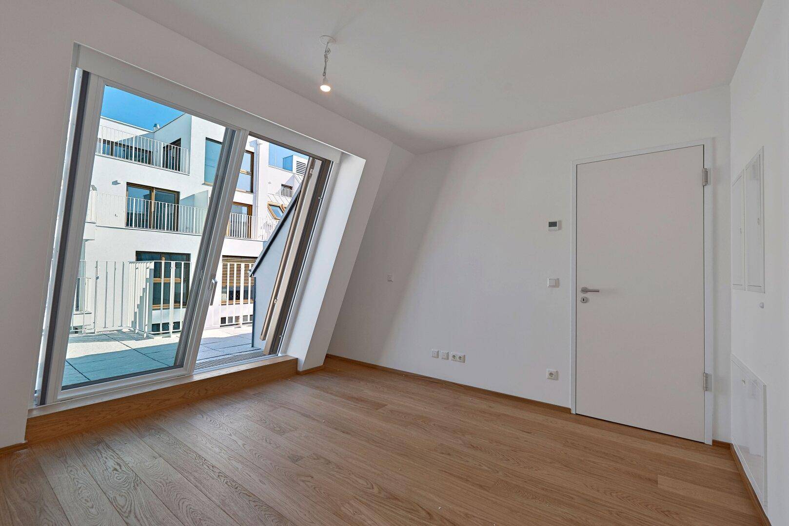 Geräumiges Zimmer mit Holzboden und großem Fenster, das den Blick auf moderne Architektur freigibt.