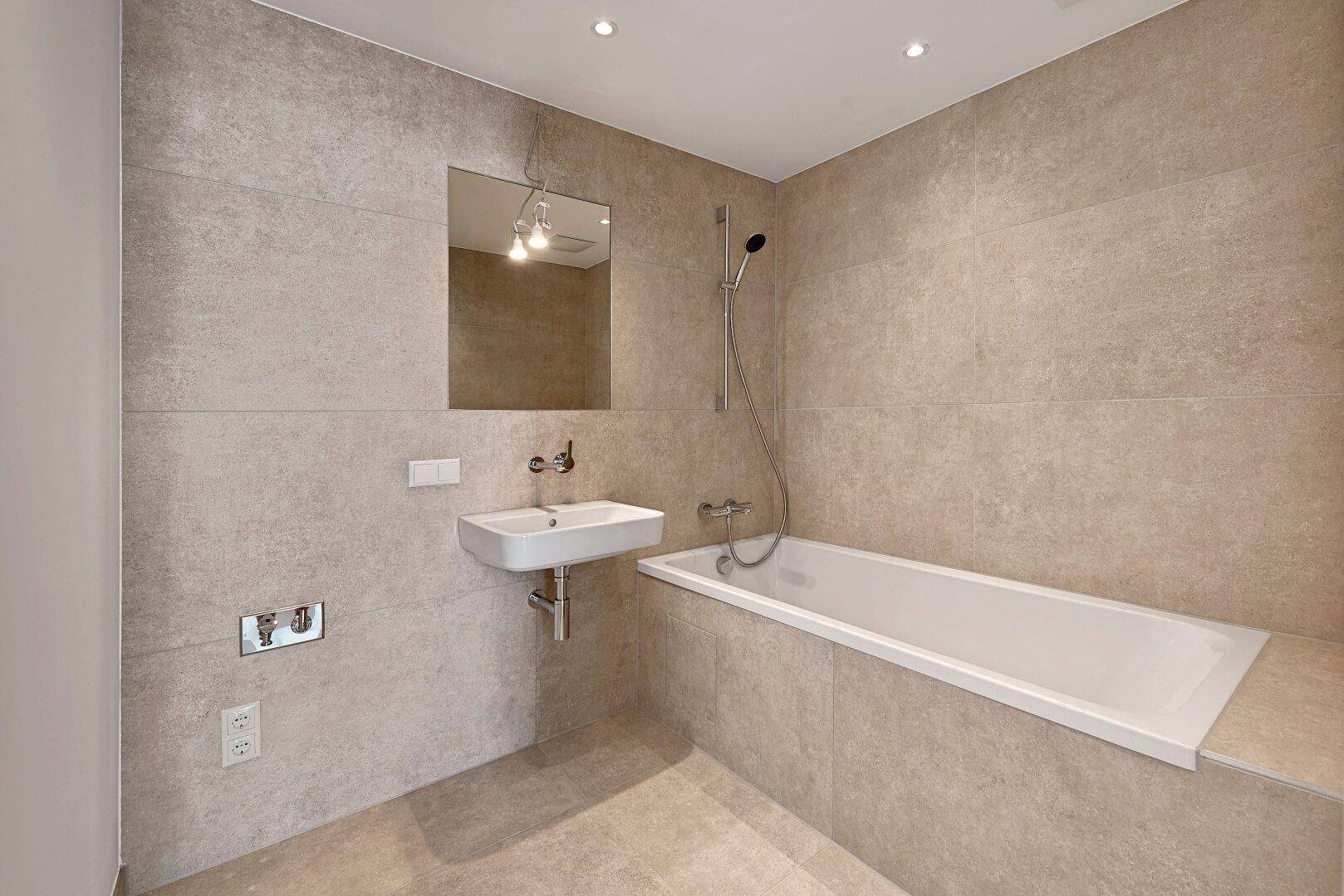 Modernes Badezimmer mit großen grauen Fliesen, Badewanne und minimalistischem Einzelwaschbecken.