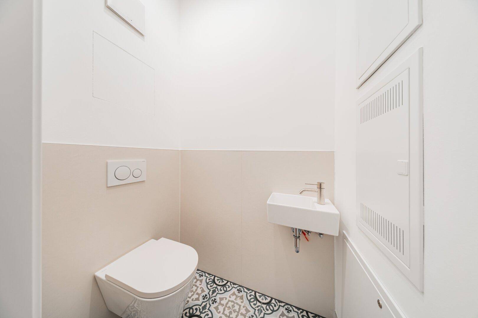 Modernes Gäste-WC mit wandhängender Toilette, kleinem Waschbecken und gemusterten Fliesen.
