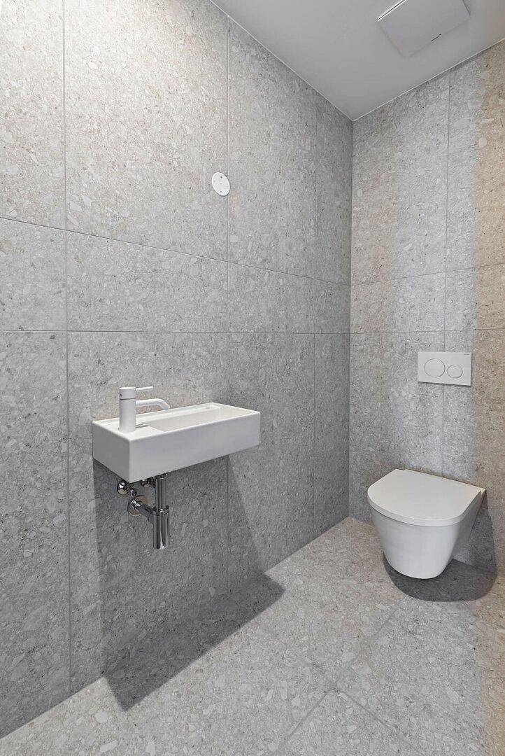 Modernes Gäste-WC mit grauen Fliesen, wandhängendem WC und kleinem Waschbecken.