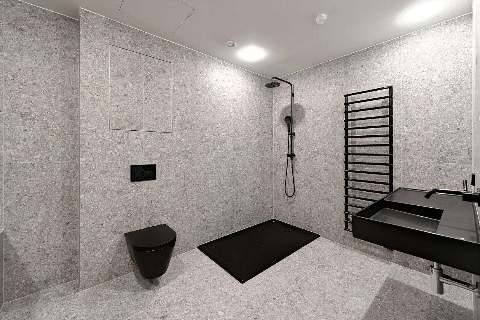 Modernes Badezimmer mit grauen Fliesen, schwarzer Dusche und wandhängendem WC.