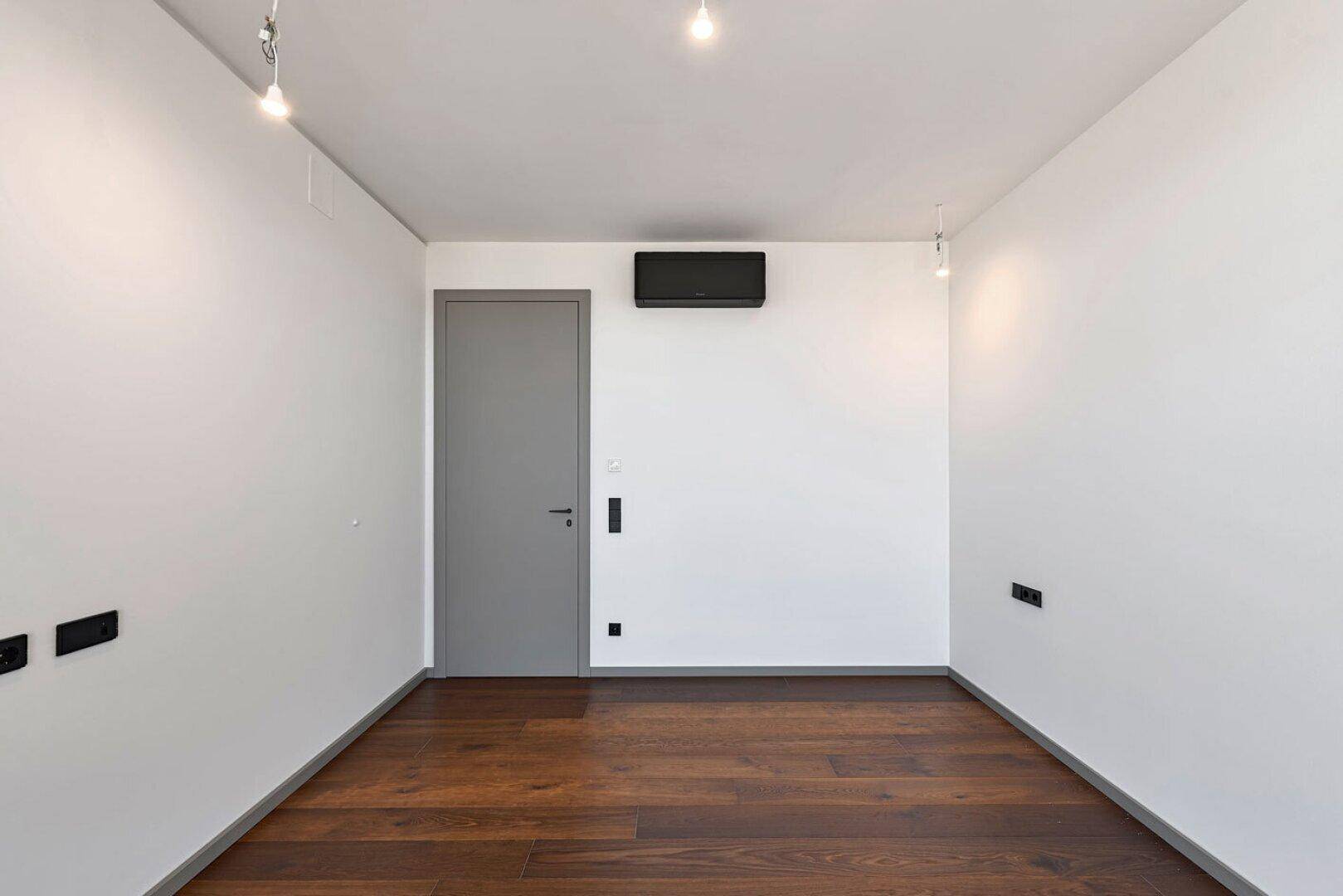 Leeres Schlafzimmer mit dunklem Parkettboden, weiß gestrichenen Wänden und Klimaanlage.