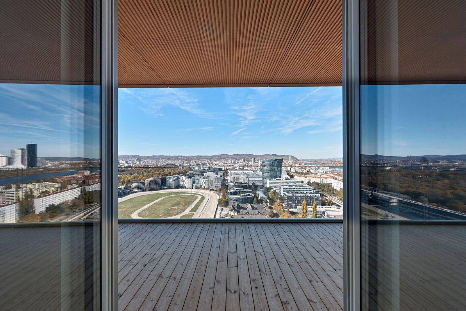 Blick von innen durch große Fenster auf die Holzterrasse und die weite Stadtlandschaft.
