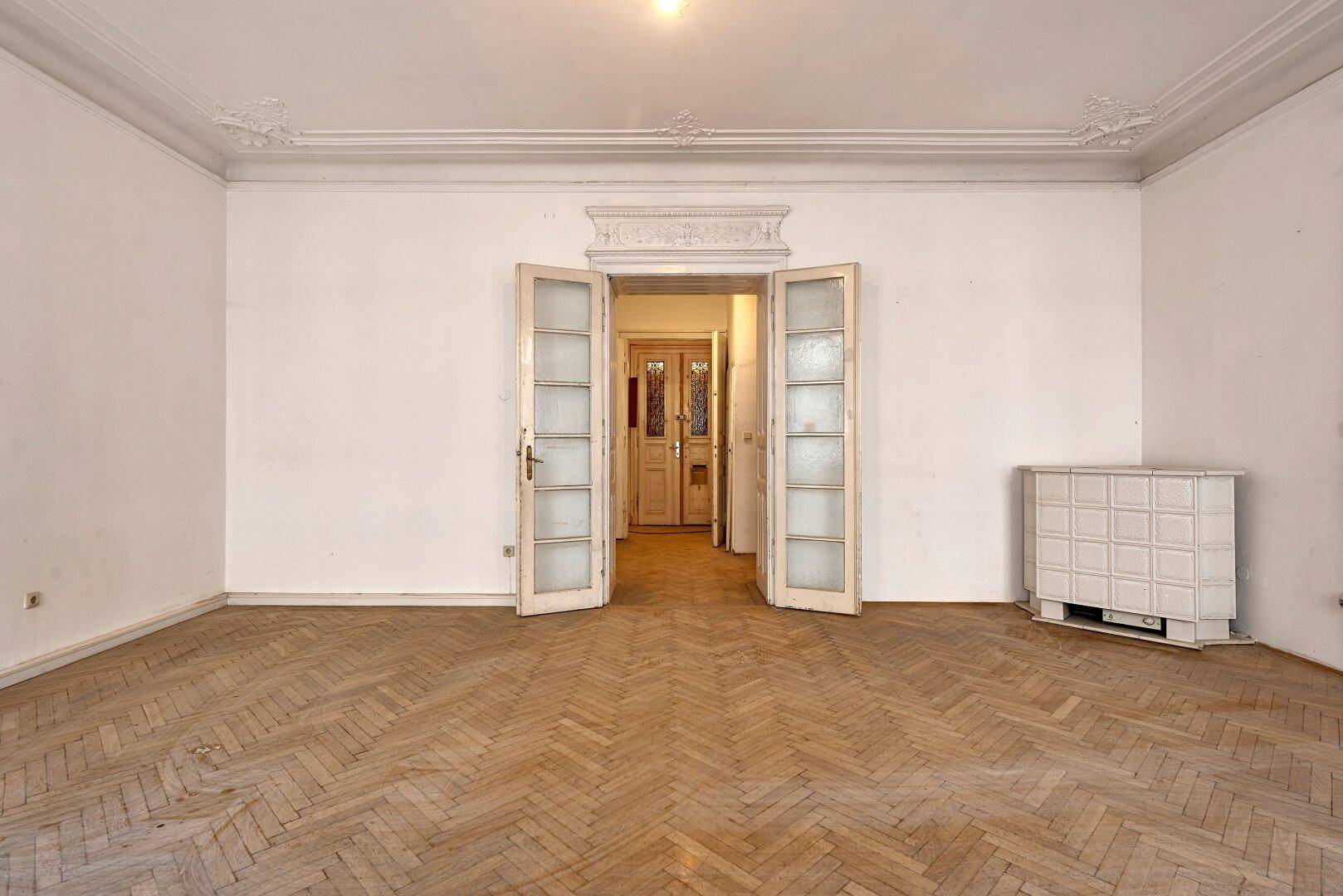 Geräumiges Zimmer mit Fischgrätparkett, hohen Decken, Stuckverzierungen und einem traditionellen Kachelofen.