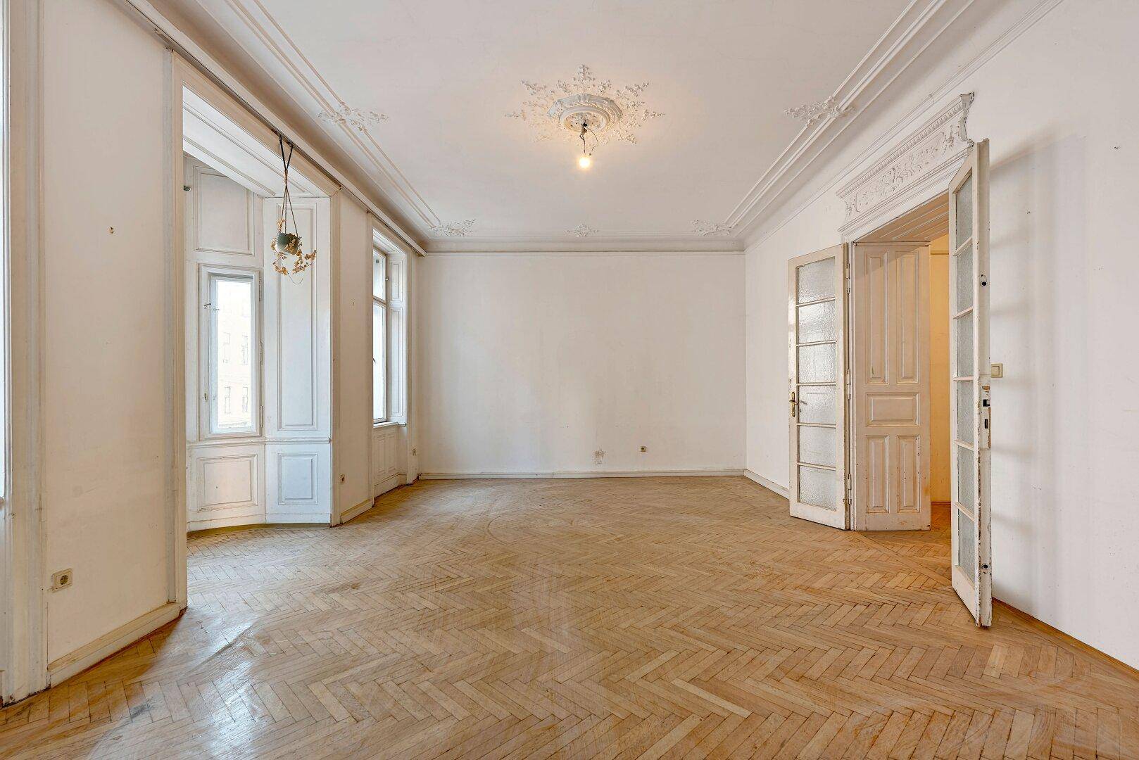 Großzügiges Wohnzimmer mit Fischgrätparkett, hohen Decken, Stuckelementen und einem Erker mit Fenstern.