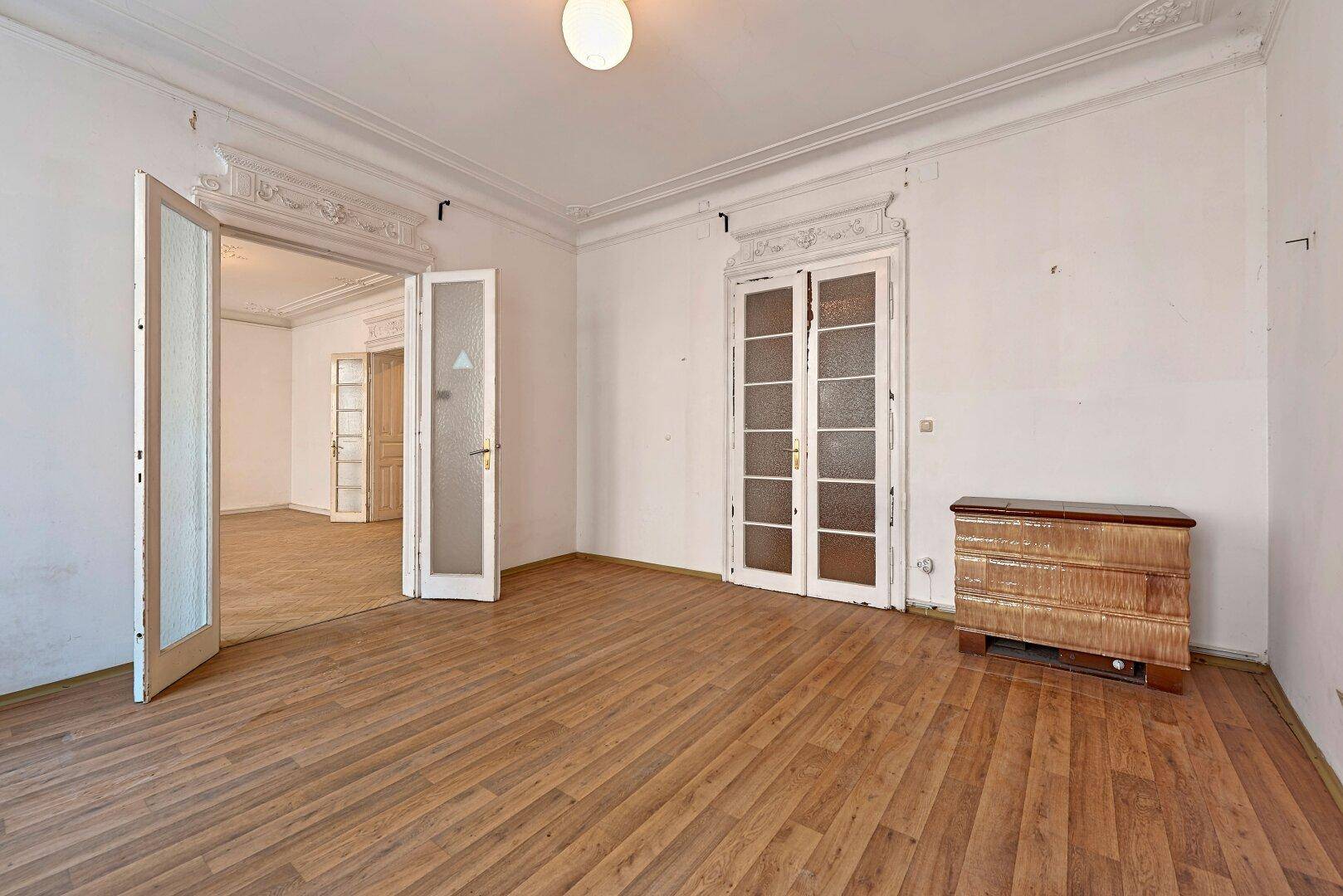 Helles Zimmer mit Laminatboden, hohen Decken, Stuckelementen und Flügeltüren zu einem weiteren Raum.