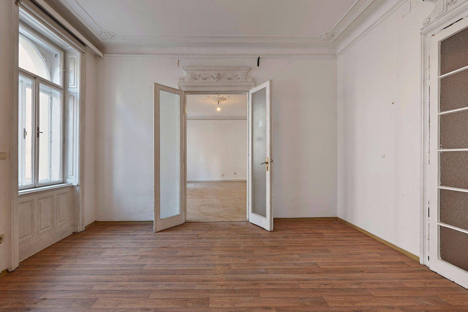 Geräumiges Zimmer mit Laminatboden, hohen Decken und Flügeltüren, die den Blick in einen weiteren Raum freigeben.
