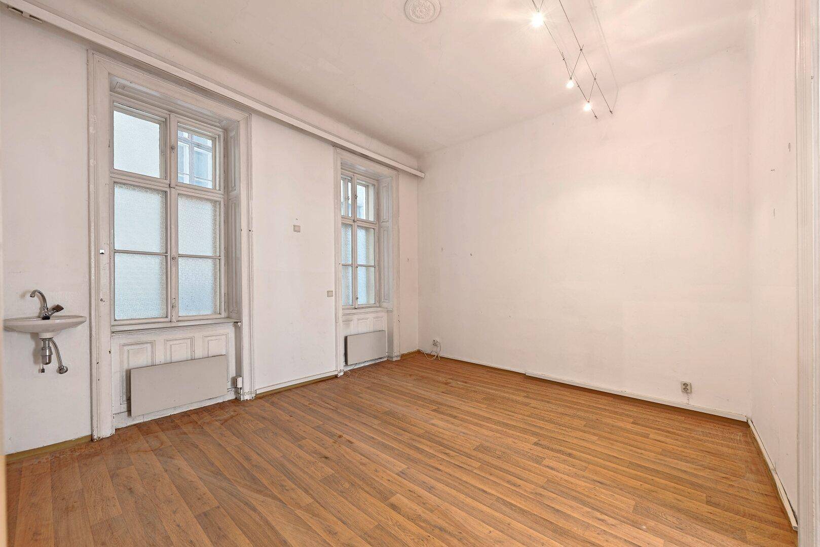 Einfacher Raum mit Holzfußboden, zwei Fenstern und einem kleinen Waschbecken an der Wand.