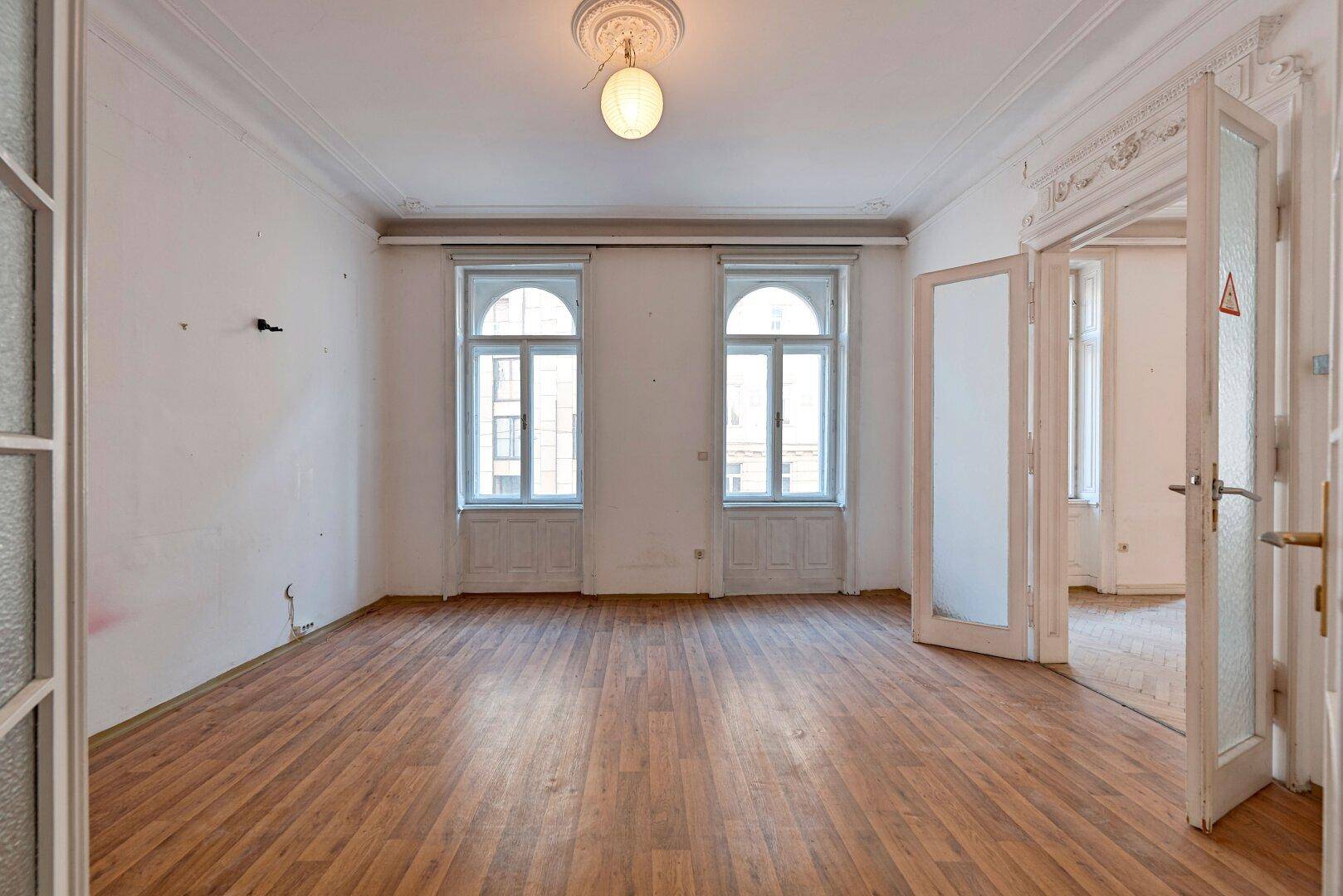 Helles Zimmer mit Laminatboden, hohen Decken, Stuckverzierungen und zwei großen Bogenfenstern.
