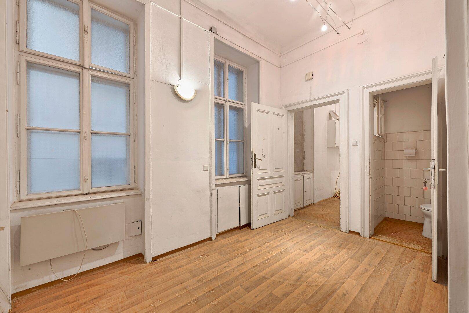 Leerer Raum mit Holzfußboden, zwei Fenstern und einem Zugang zu einem Badezimmer.