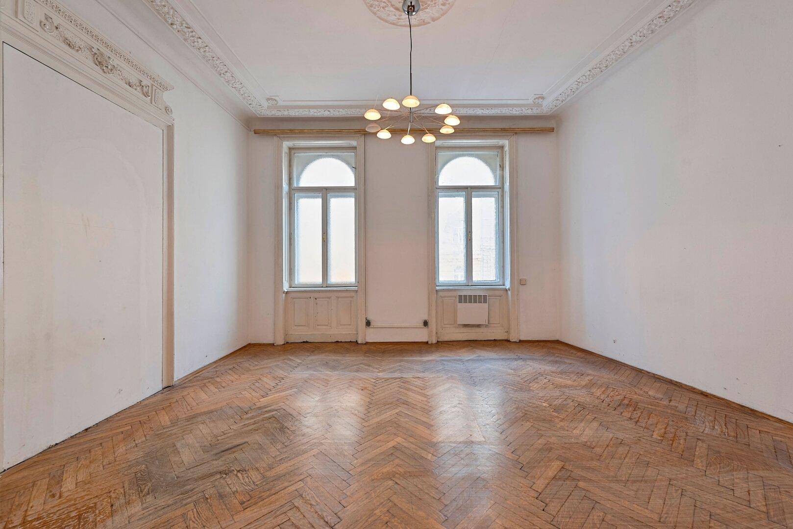 Geräumiges Zimmer mit Fischgrätparkett, hohen Decken, Stuckverzierungen und einem modernen Kronleuchter.