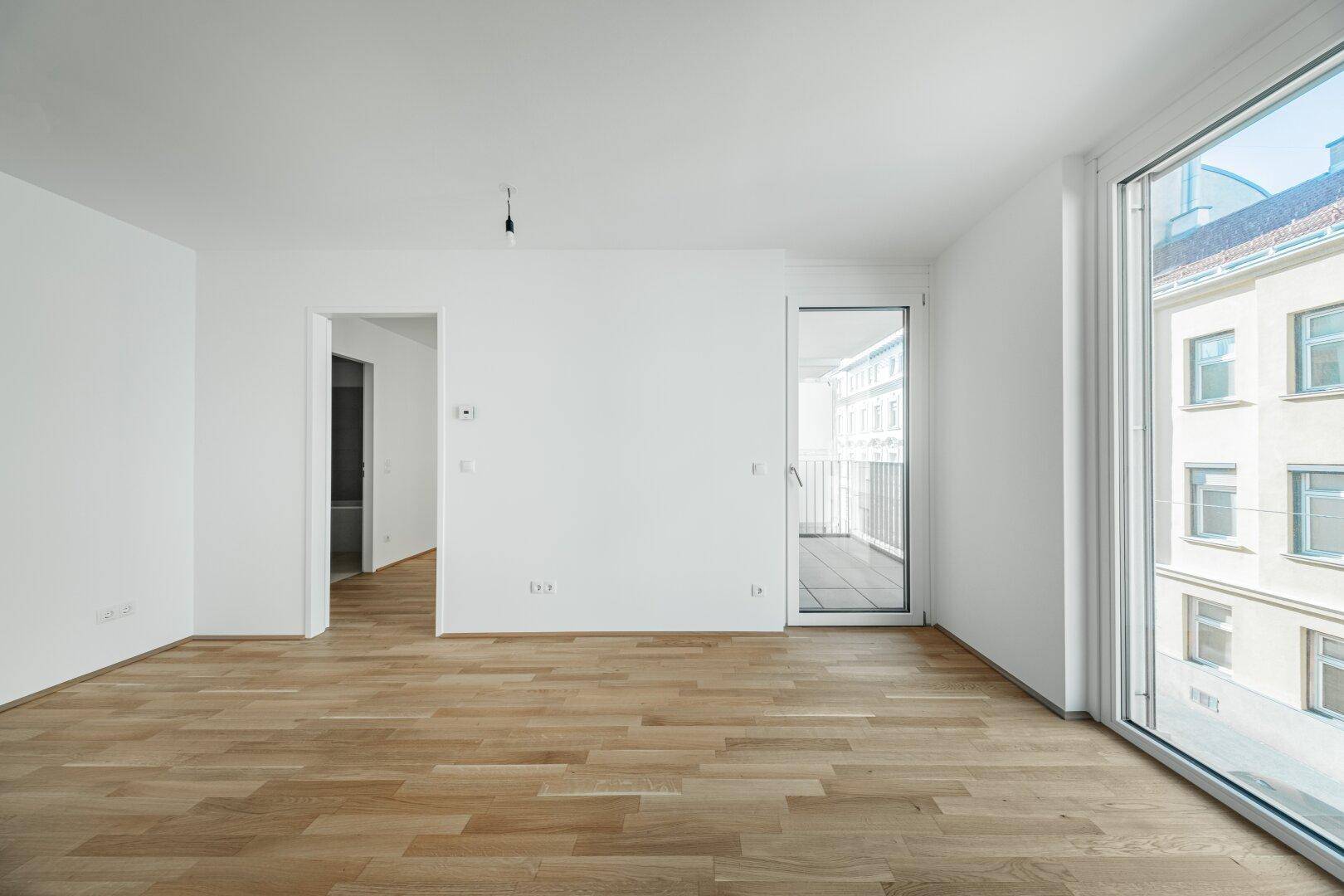 Geräumiger, leerer Raum mit hellem Parkettboden und großem Fenster, das Zugang zu einem Balkon bietet.