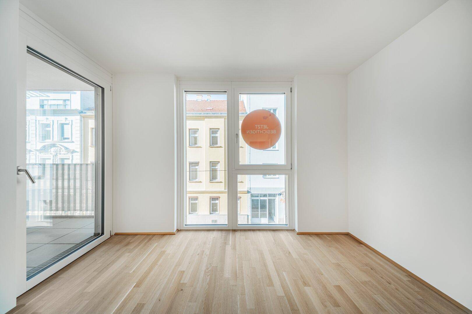 Leerer Raum mit hellem Parkettboden und großem Fenster, das viel Tageslicht hereinlässt.