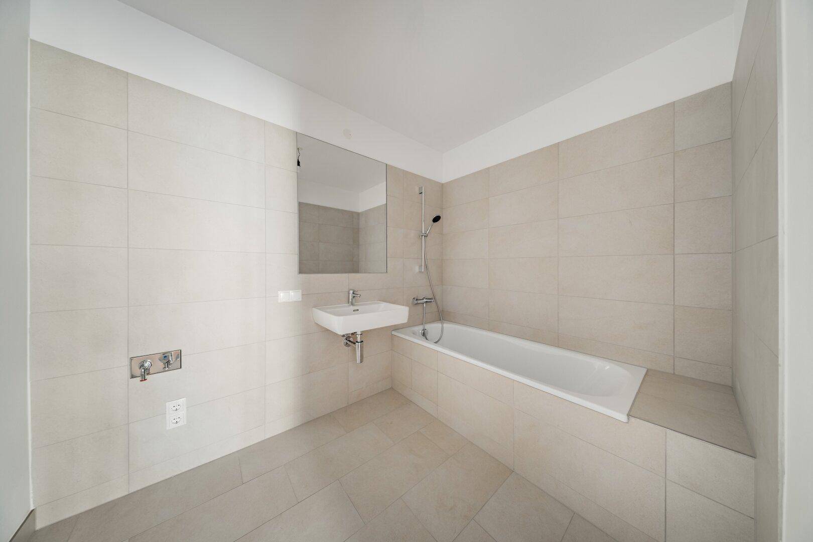 Helles Badezimmer mit Badewanne, Waschbecken und beigefarbenen Fliesen an Wänden und Boden.